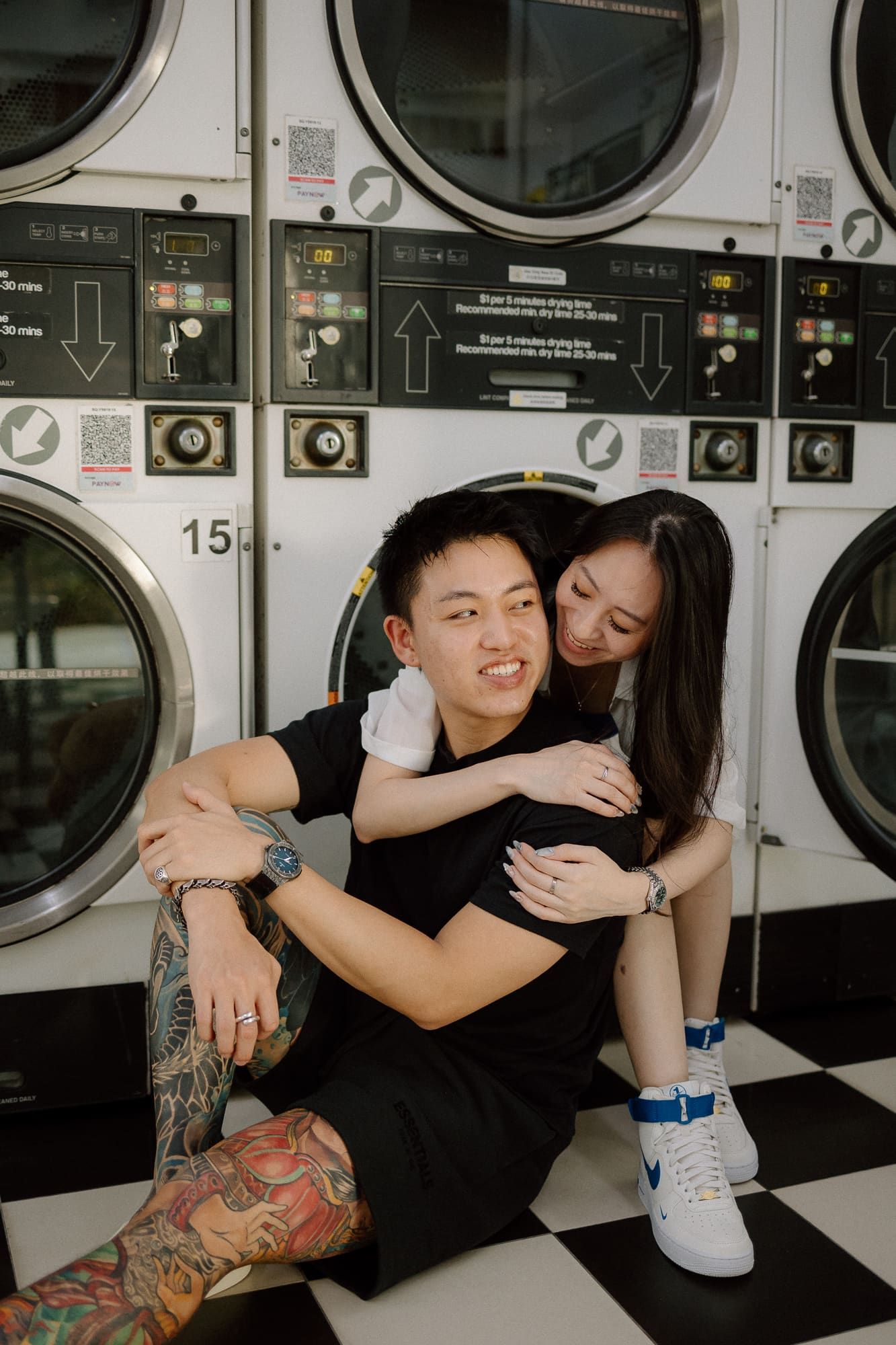 A couple shares an intimate moment while sitting on the floor of a laundromat with industrial washing machines in the background.