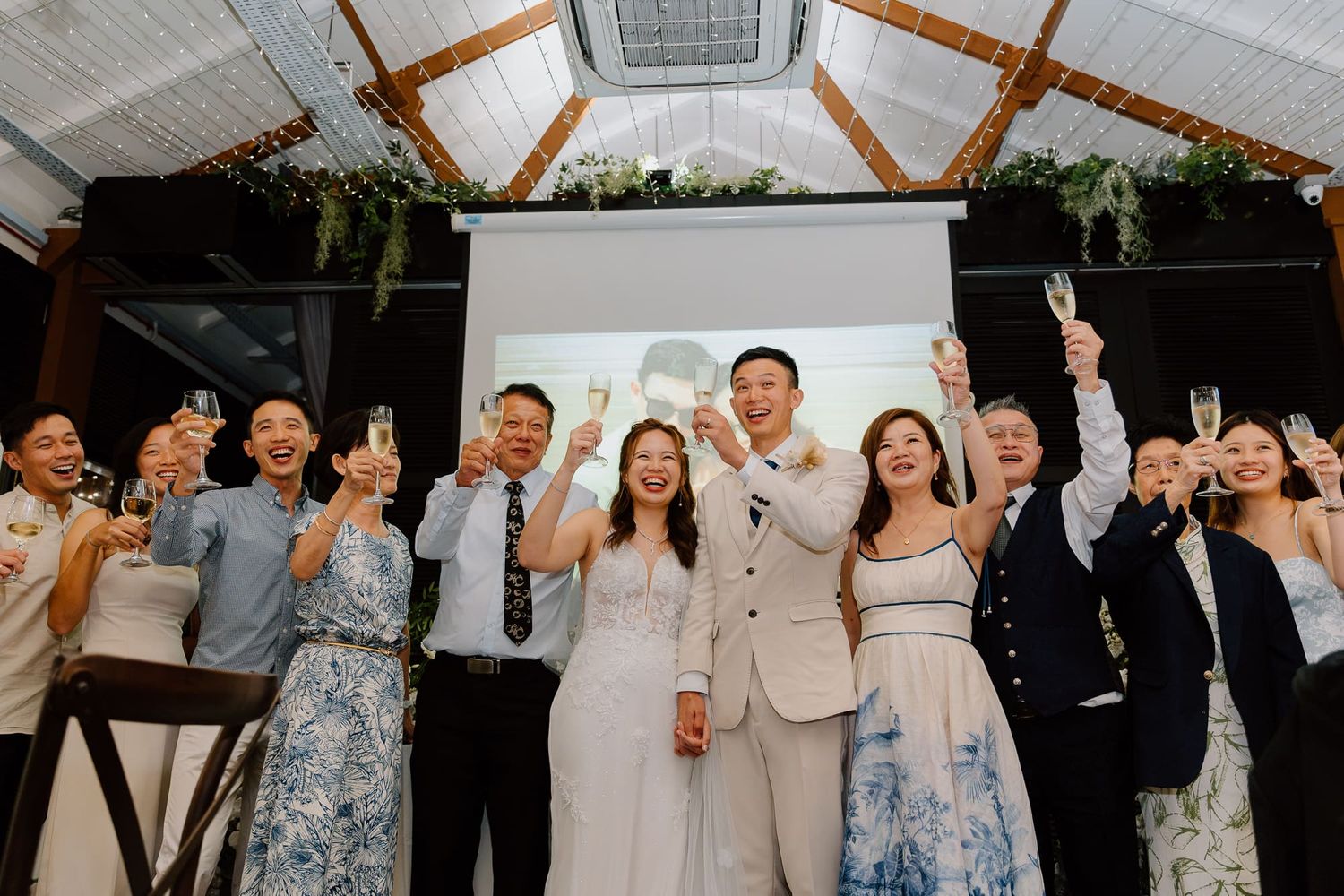 Wedding party celebrates joyfully under a glass ceiling decorated with greenery at an indoor venue.