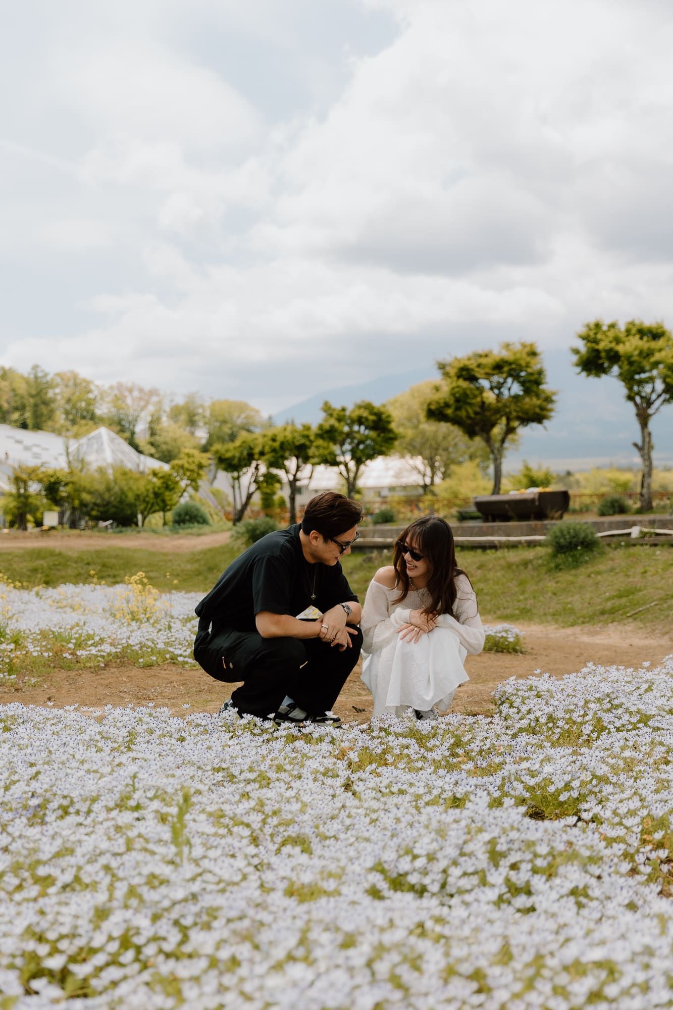 Couple crouching among blue nemophila flowers in a scenic garden with trees and cloudy sky.