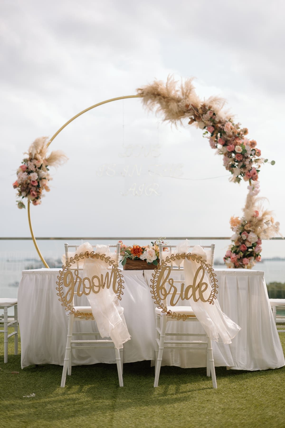 Elegant outdoor wedding setup with white chairs adorned with floral arrangements and pampas grass decor on a grassy lawn.