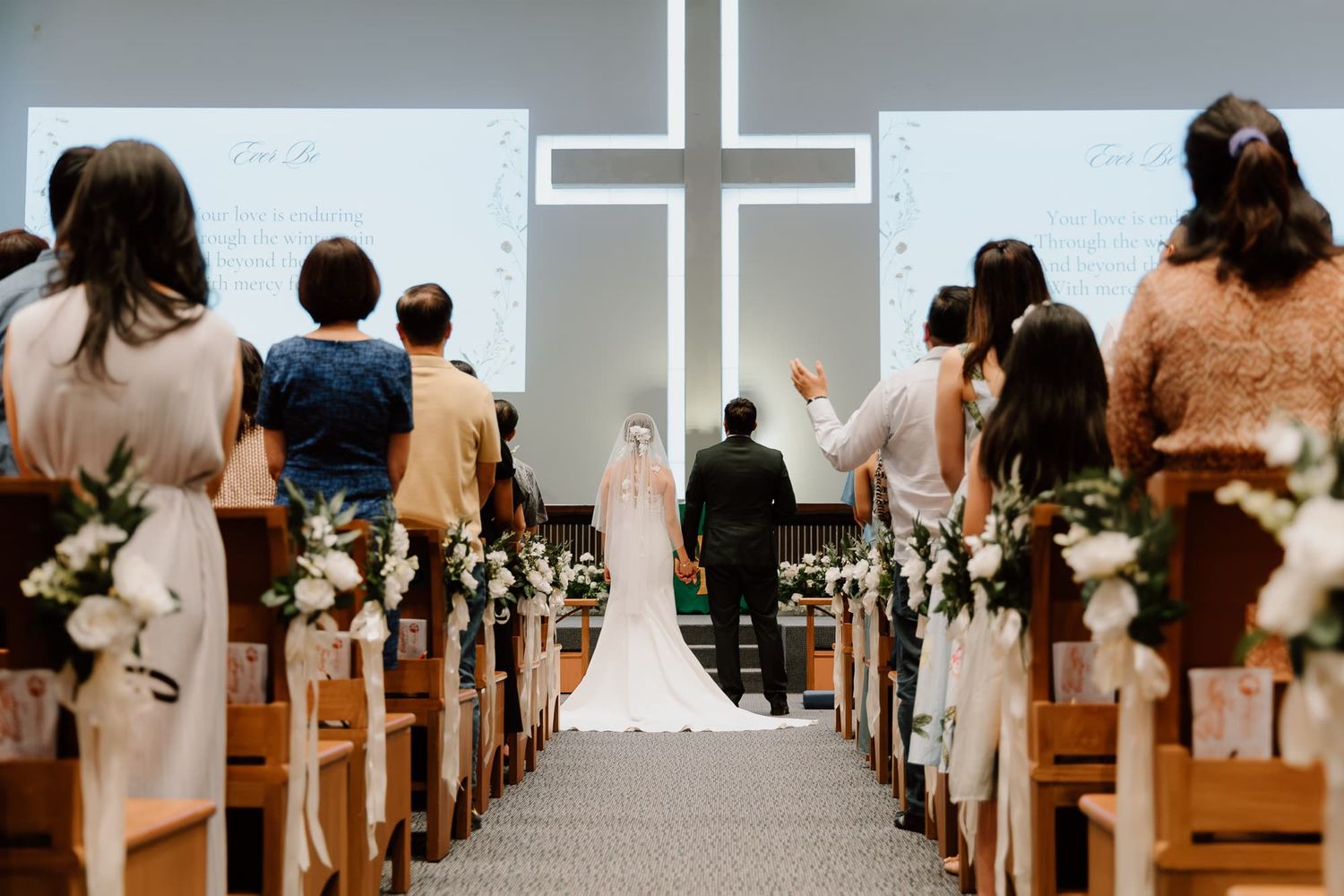 View down church aisle with illuminated cross and decorated pews during wedding ceremony.