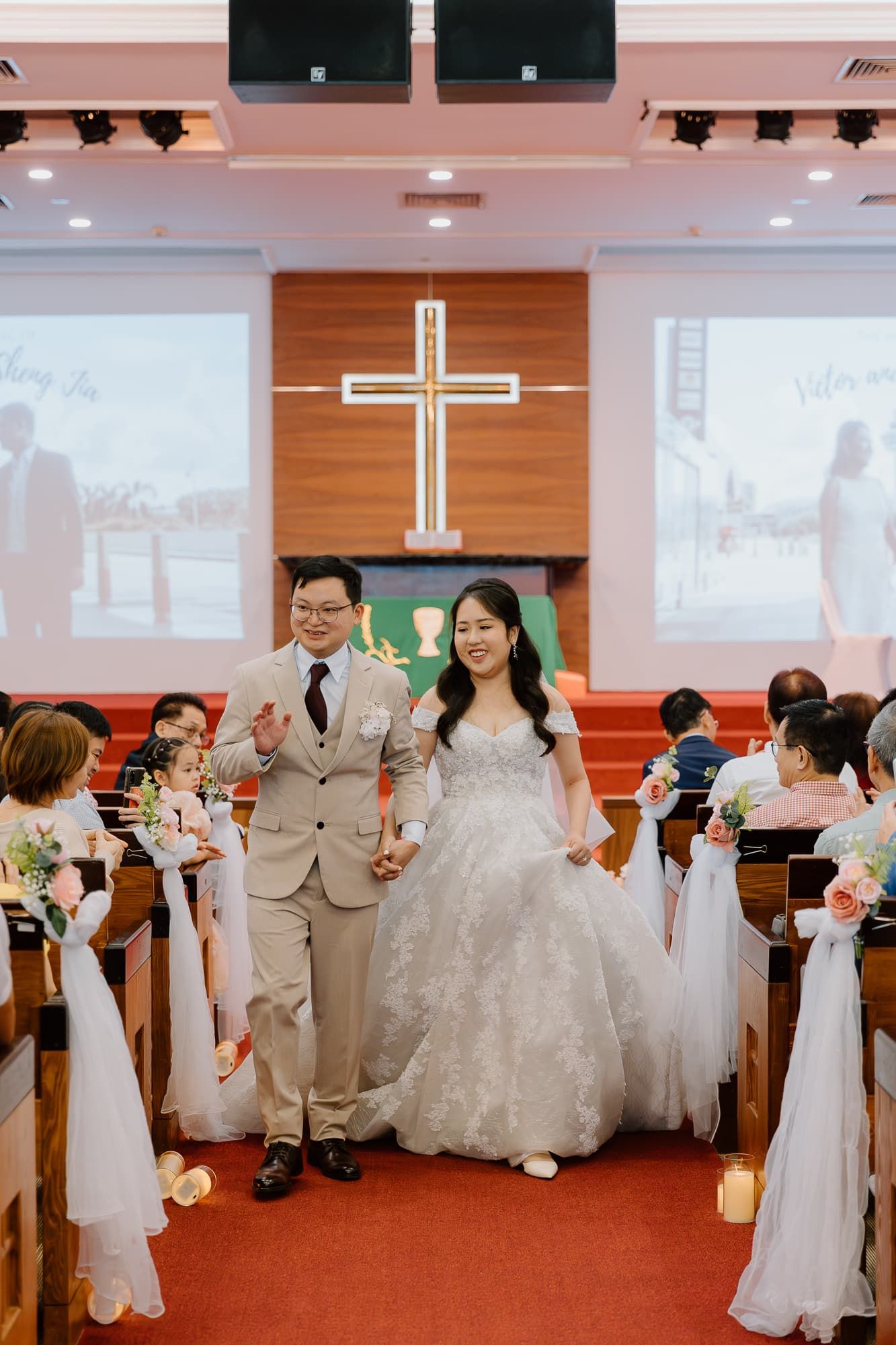 Newlyweds walk down the aisle in a church sanctuary decorated with white bows and a wooden cross on stage.
