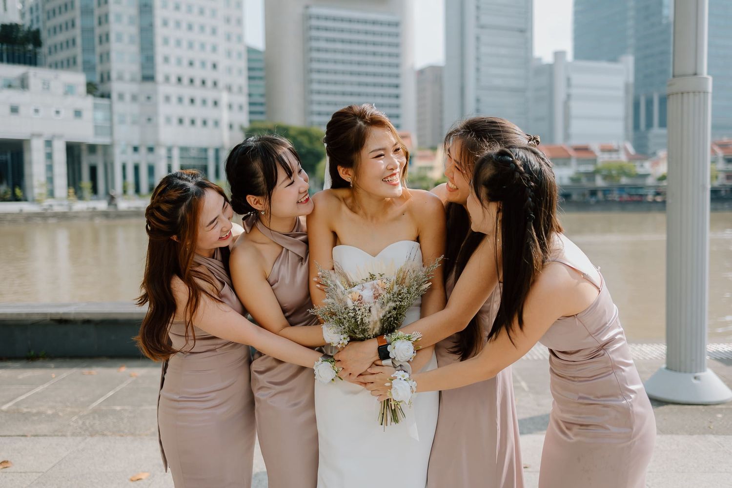 A bride and her bridesmaids share a joyful moment together on a terrace overlooking an urban skyline.