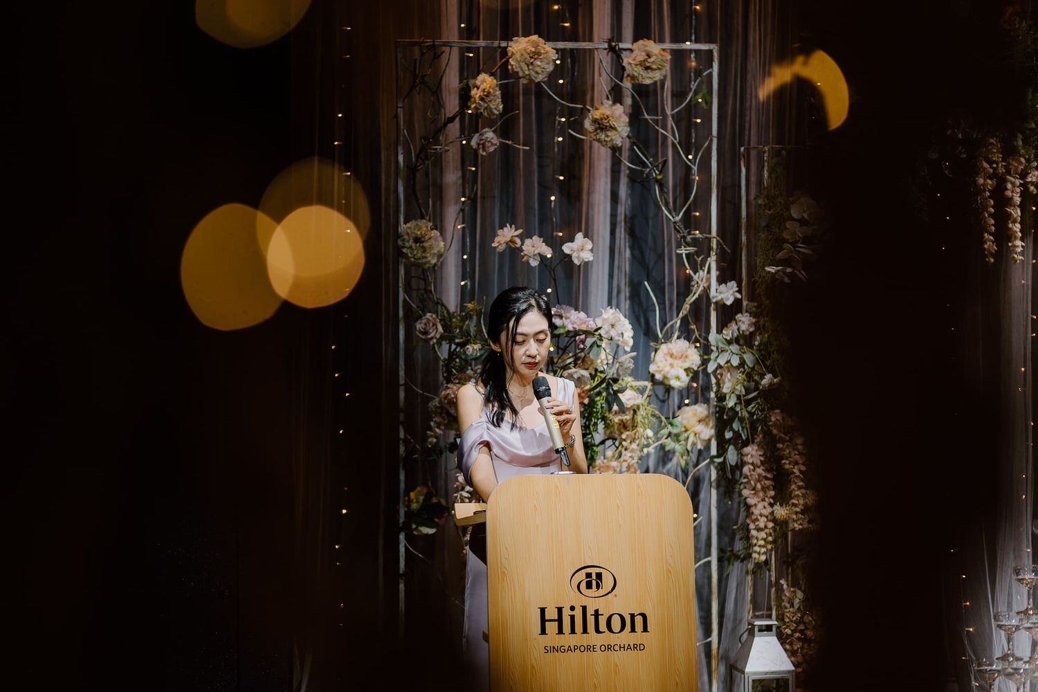 Illuminated Hilton hotel logo sign glows warmly in dimly lit interior with decorative lights and floral arrangements.