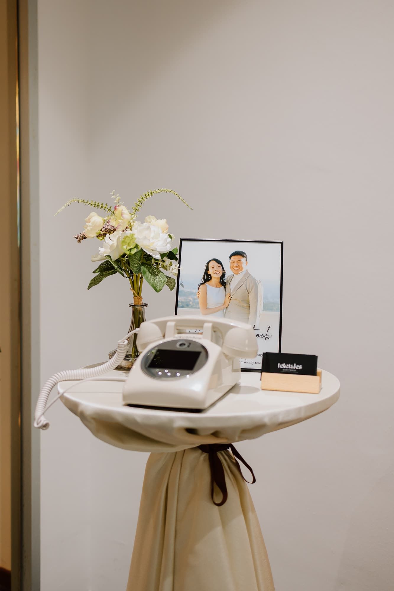 Vintage white telephone and floral arrangement on draped table with framed photograph.