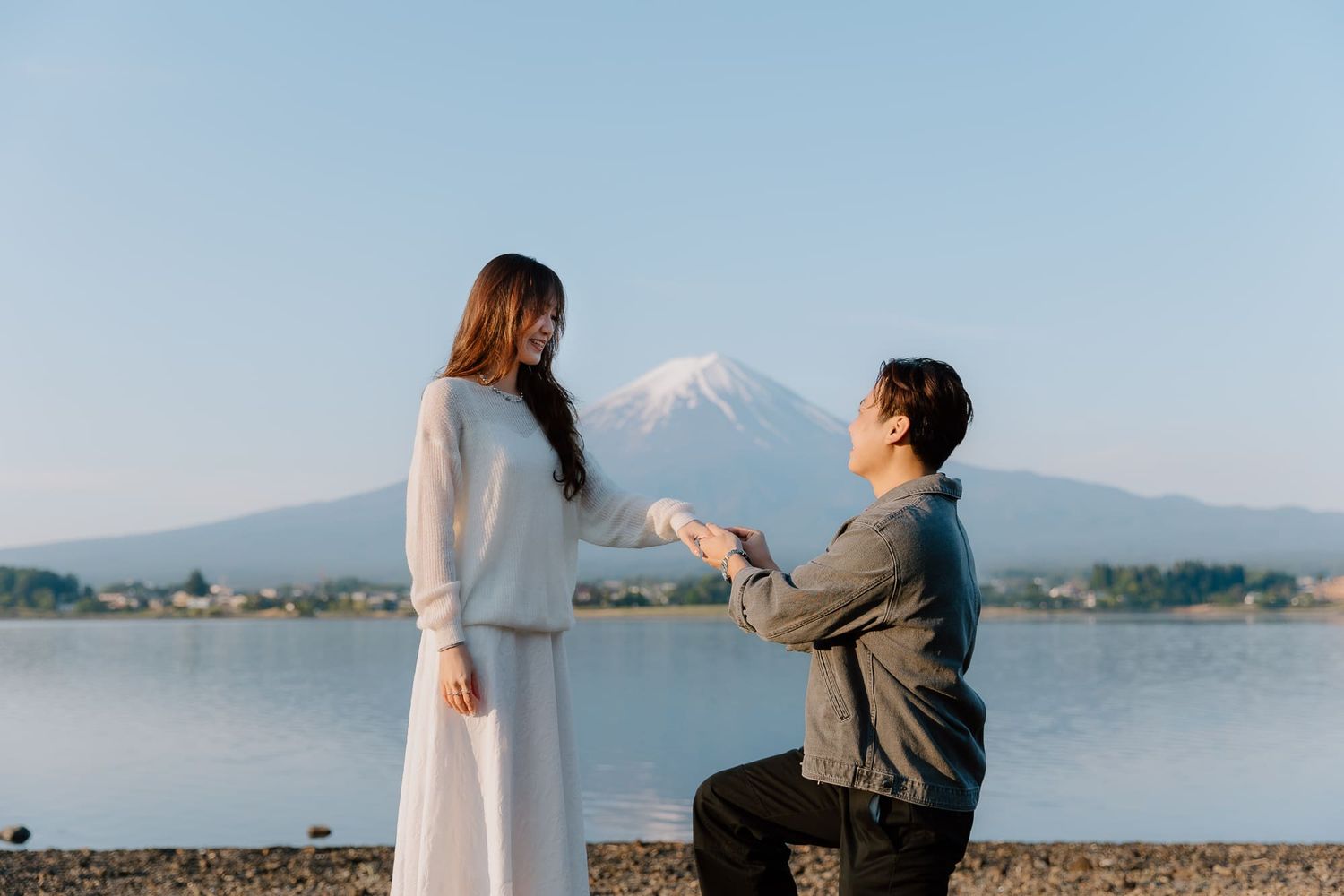 Man proposes to woman on one knee by a lake with Mount Fuji in the background on a clear day.