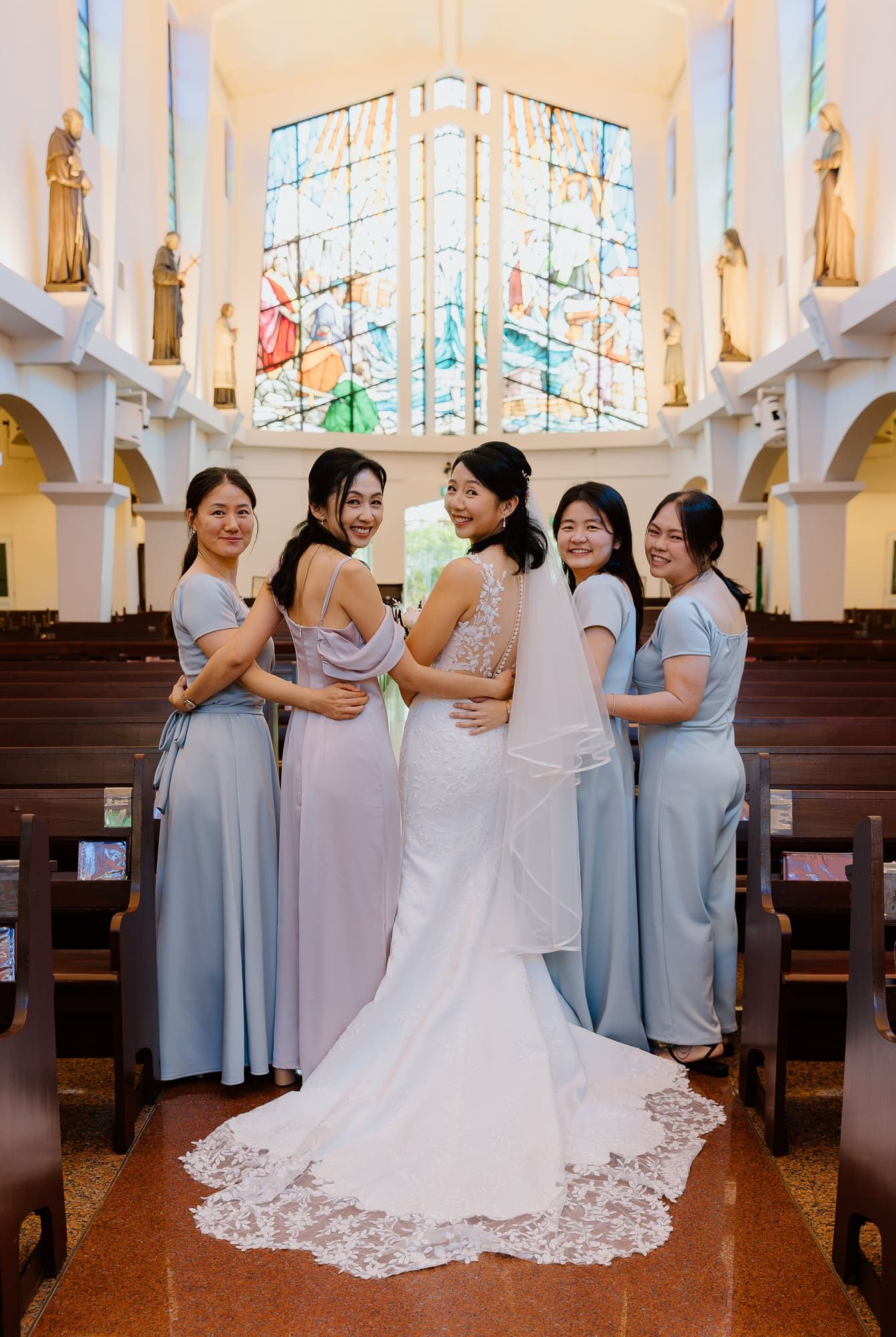A bridal party in pale blue bridesmaid dresses gathers joyfully around a bride in a white lace wedding gown inside a church.
