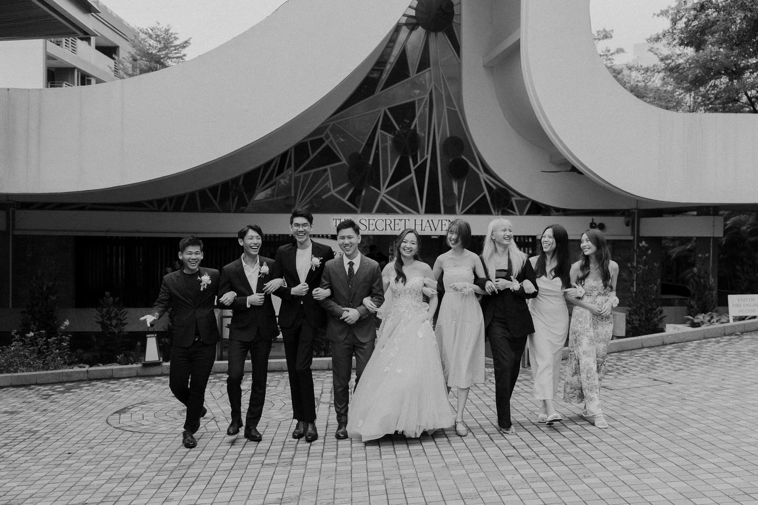 Black and white photo of wedding party walking together outside modern architectural venue.