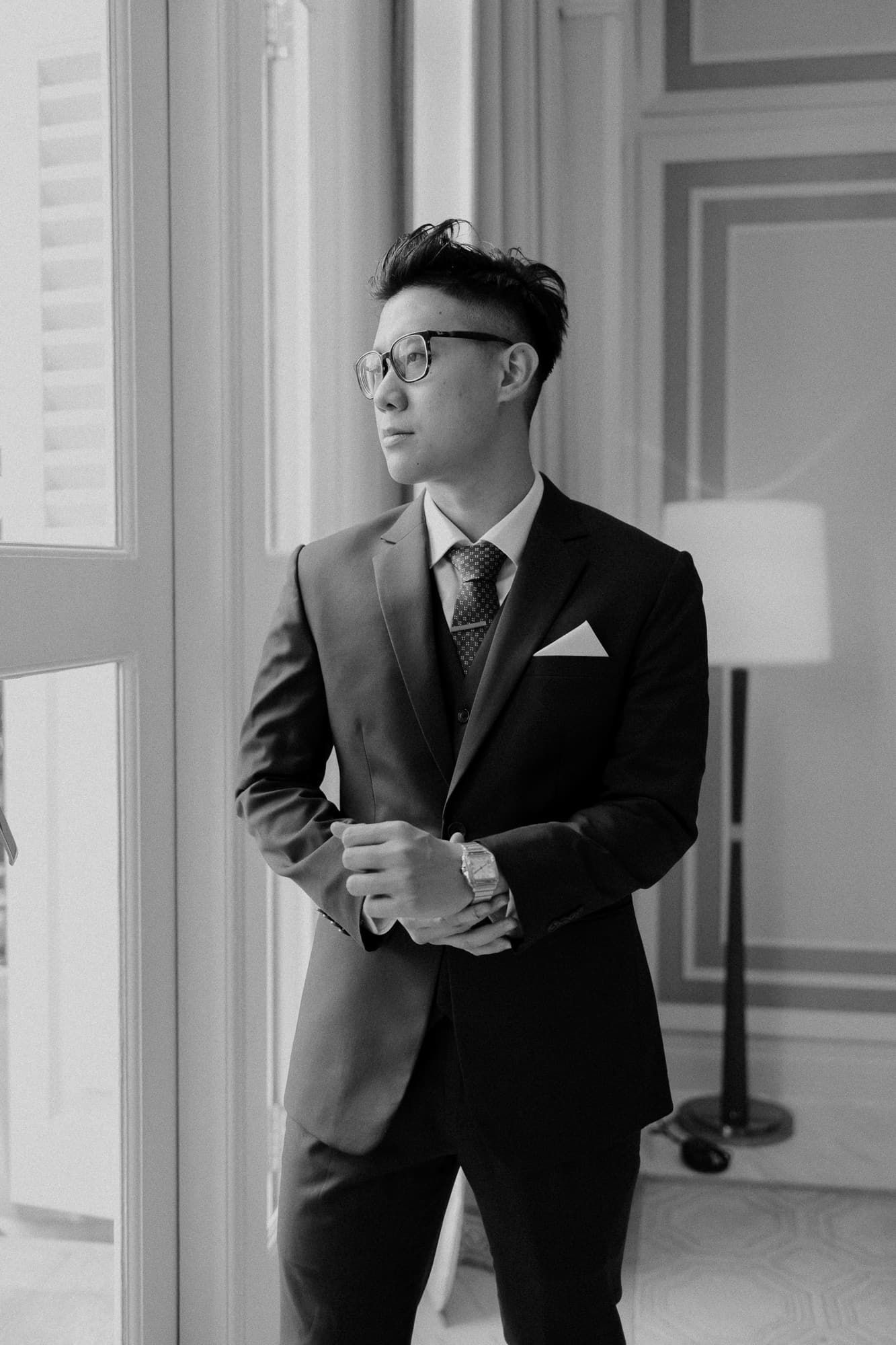 A formal portrait showing a suited figure standing by a window in black and white.