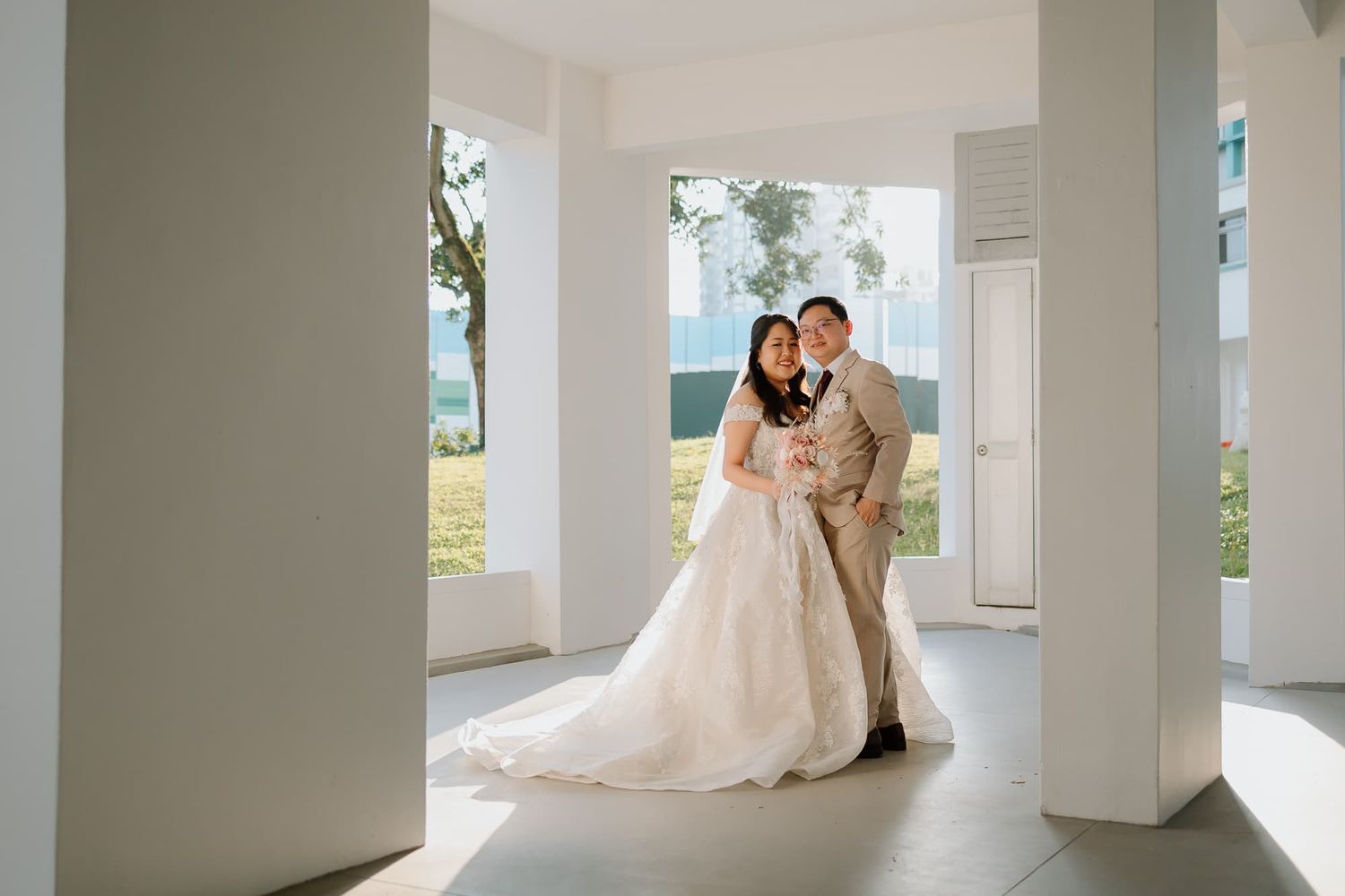 A wedding couple poses elegantly between white columns on a bright porch.