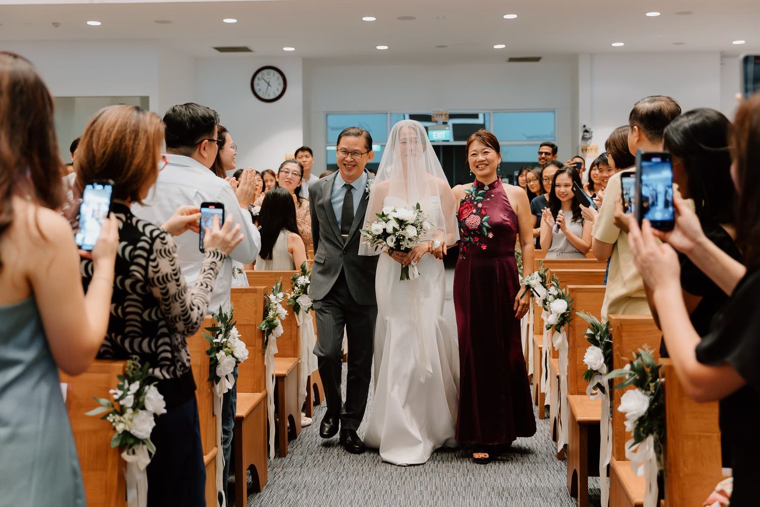 Bride in white dress walks down church aisle with parents while guests take photos.