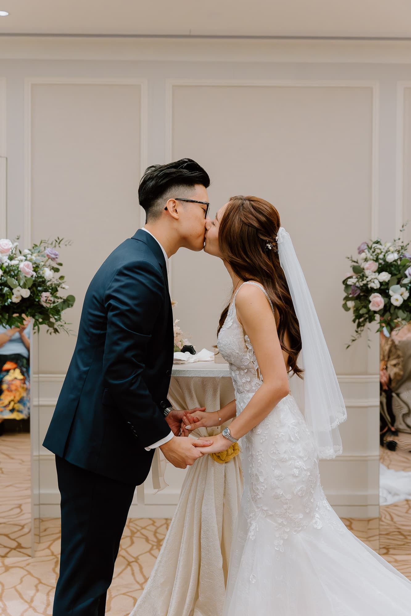 Romantic first kiss as newlyweds surrounded by elegant floral arrangements at indoor ceremony.
