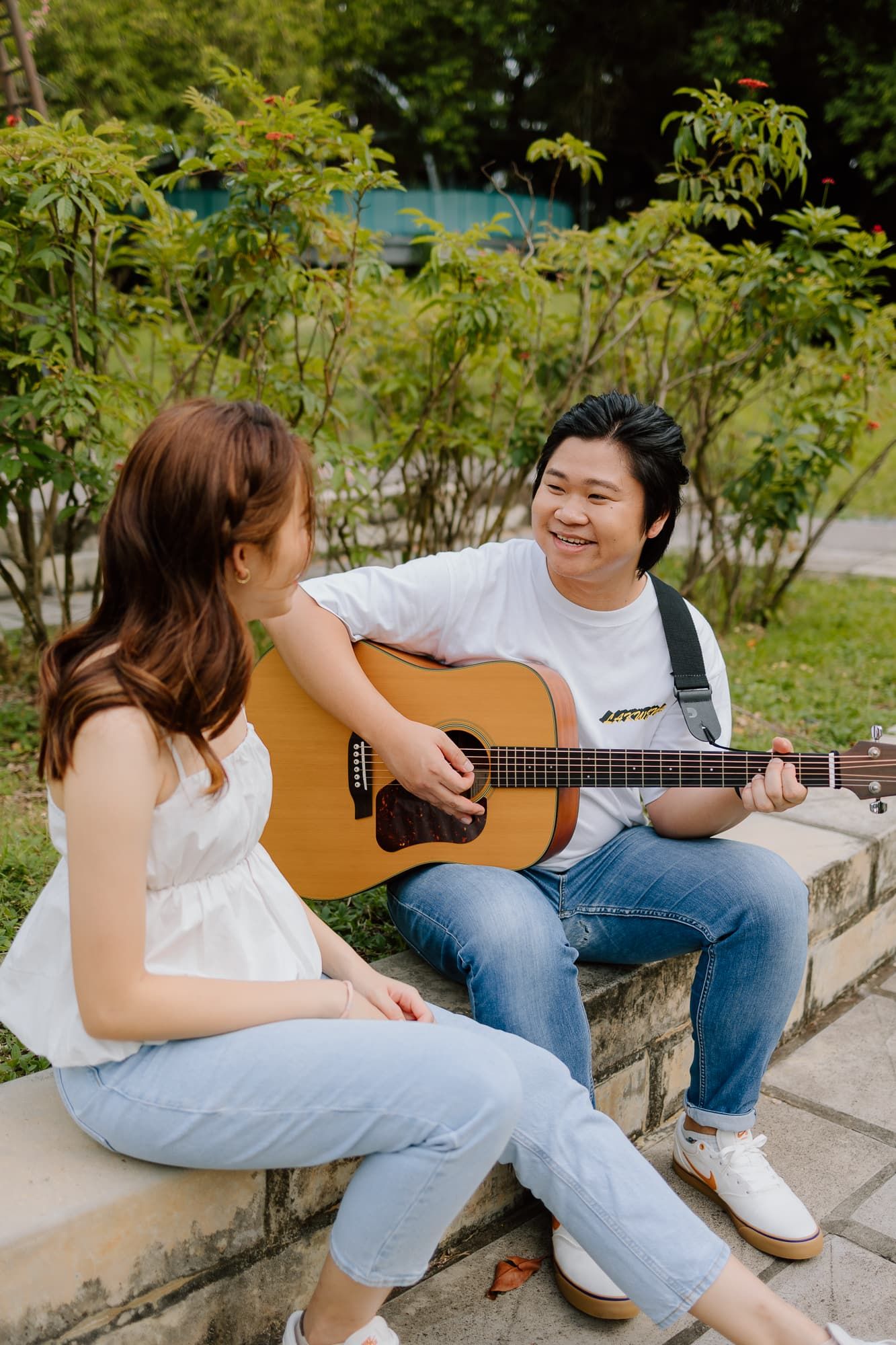 Two people sitting outdoors as one plays acoustic guitar on stone wall surrounded by greenery.