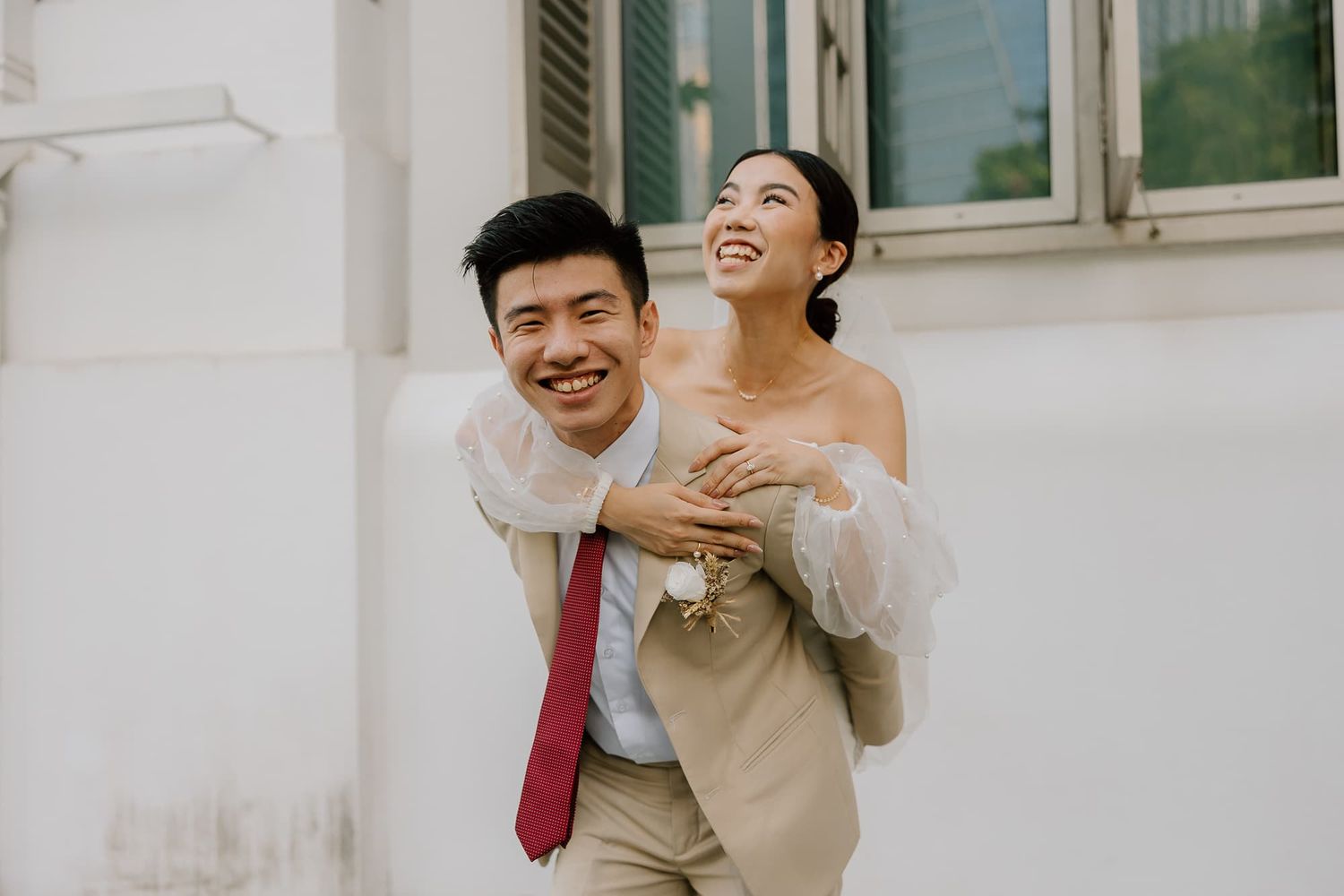 A couple shares a playful moment in their wedding attire against a white wall.