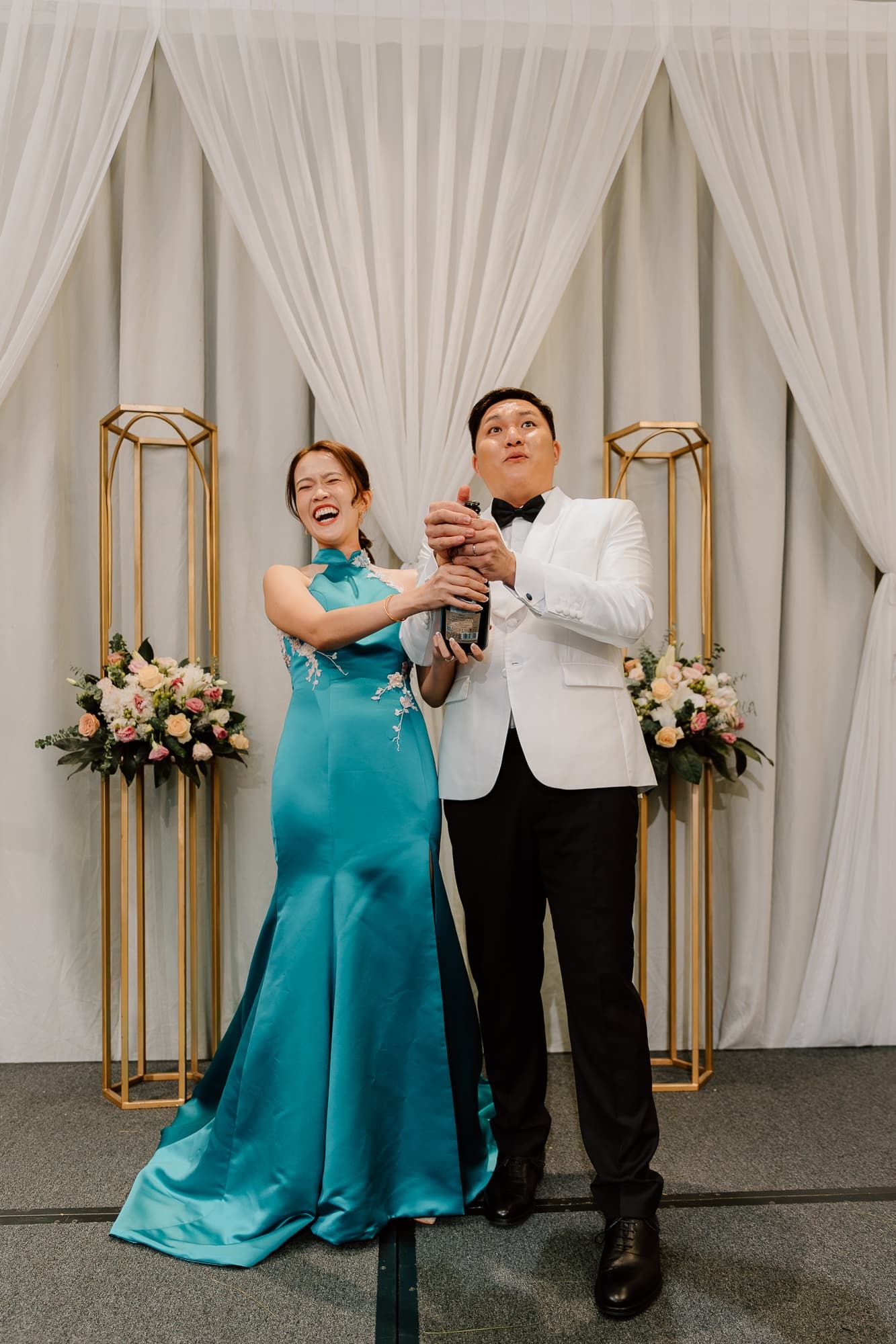 Couple in formal attire shares a joyful moment against white drapes and gold decor.