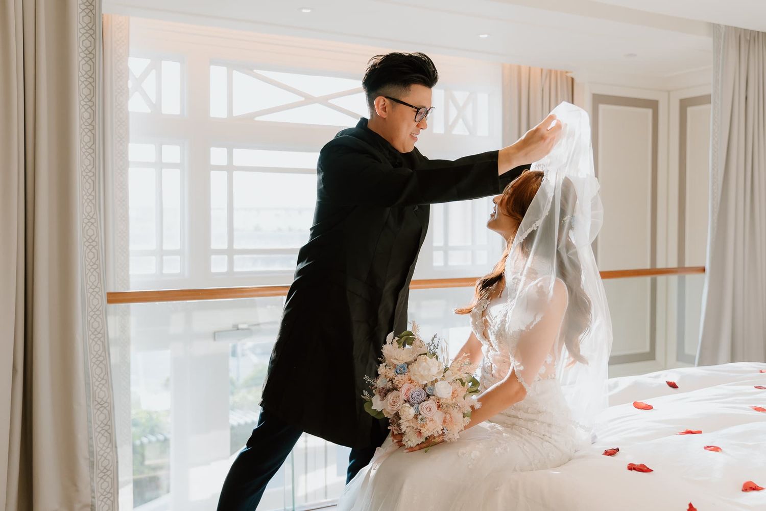 Playful wedding couple dances together holding floral bouquet near large windows.