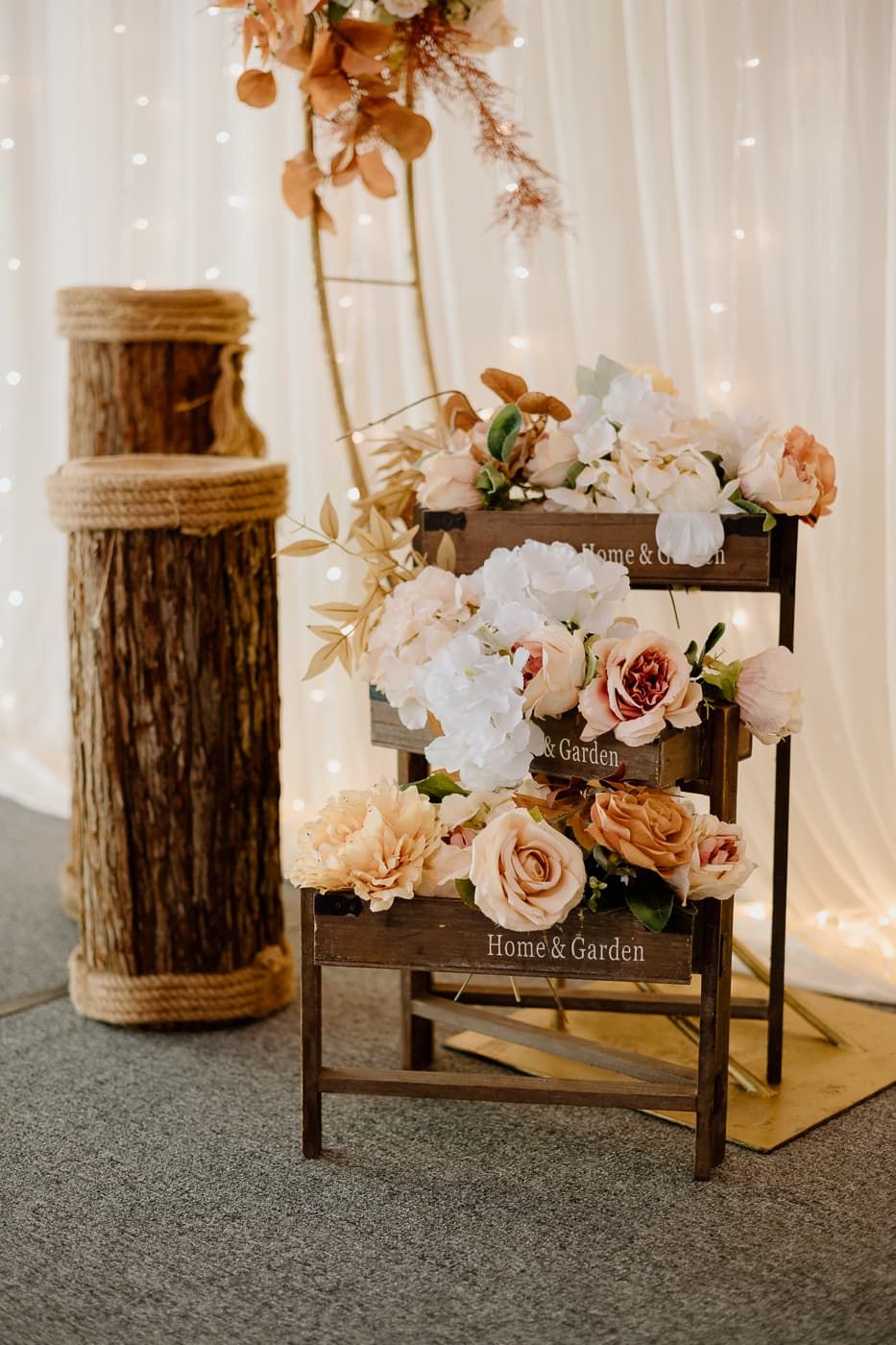 Rustic floral arrangement with roses in wooden display stand against string light backdrop.