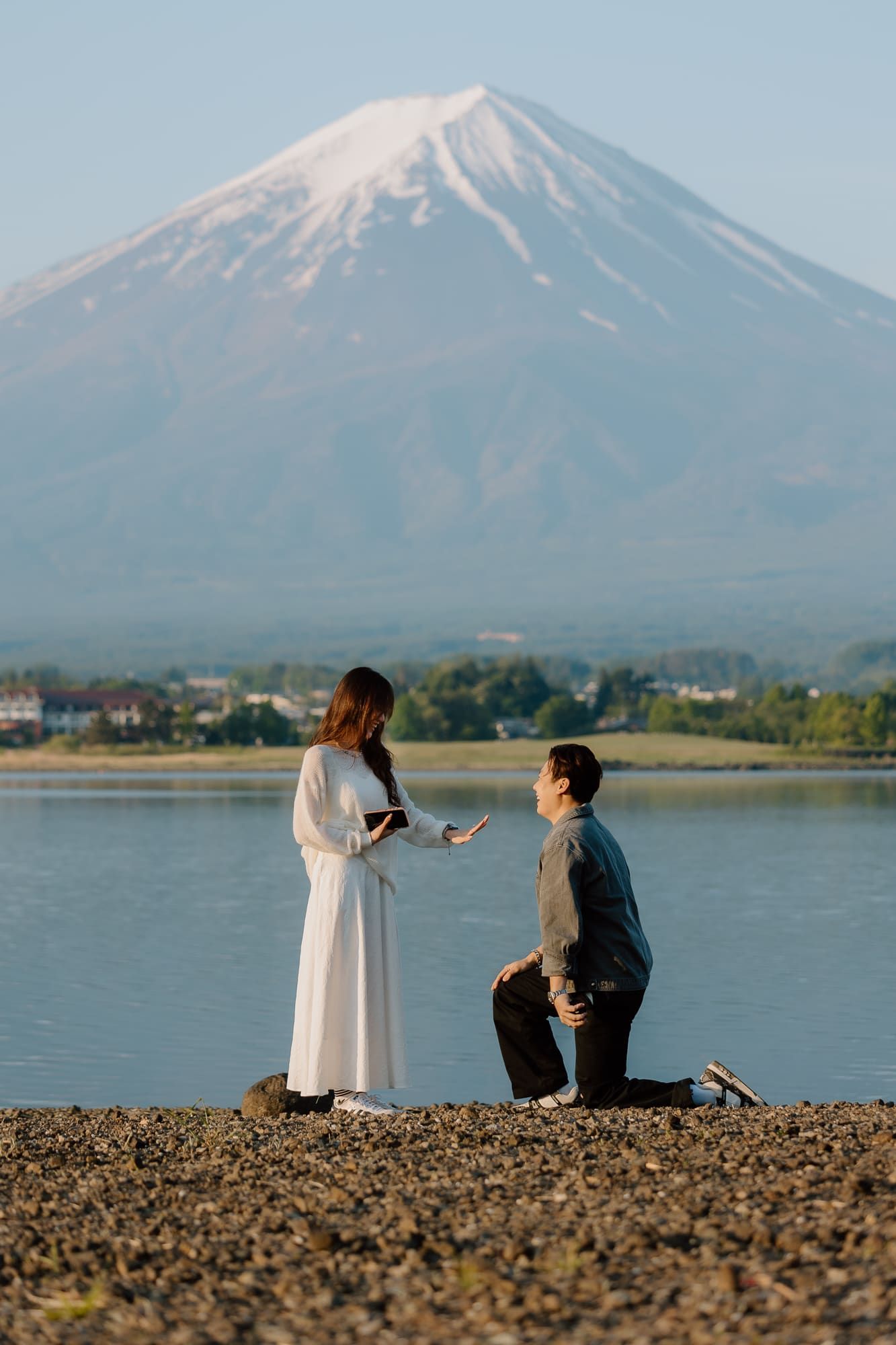 Man proposing to woman in white dress by lake with Mount Fuji in background on rocky shore.