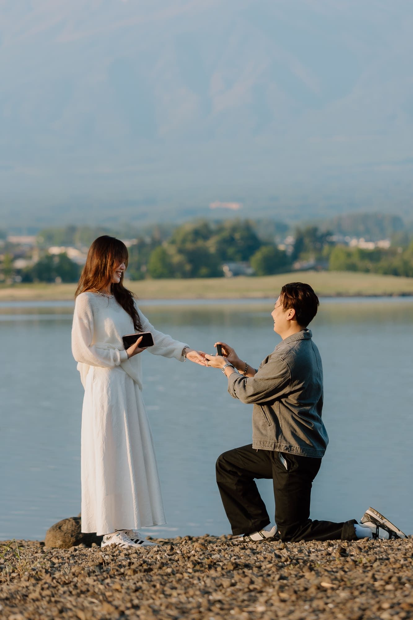 Man proposing on one knee to woman in white dress by a scenic lake with mountains in the background.