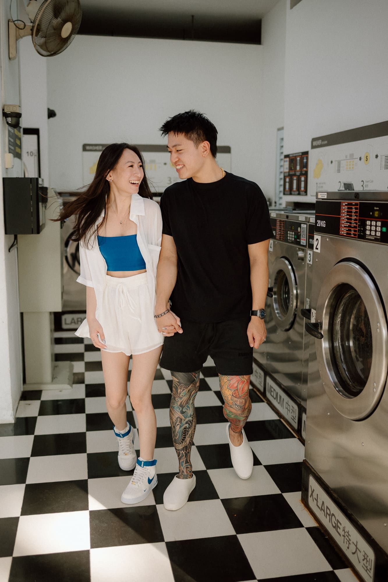 A couple holding hands while walking through a laundromat with black and white checkered floors.