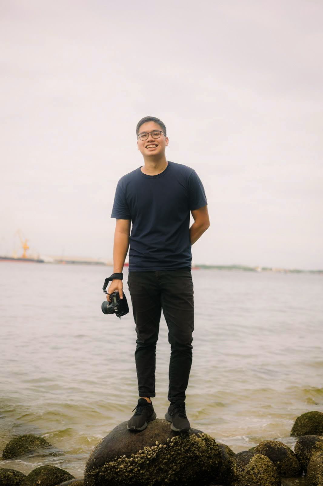 A photographer in black attire stands on rocky shore overlooking water and distant harbor views at sunset.