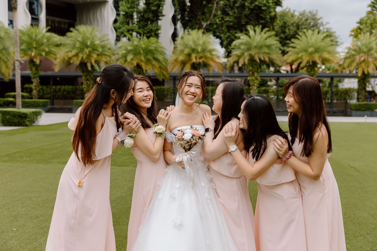 Bridesmaids in pink dresses surround bride in white gown sharing joyful moment outdoors.