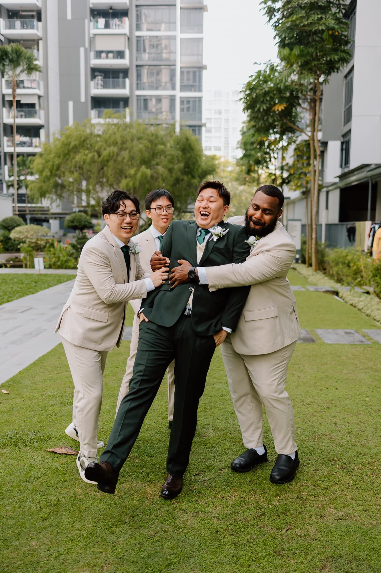 Groomsmen in light suits share a candid laughing moment together on a lawn with apartment buildings in background.