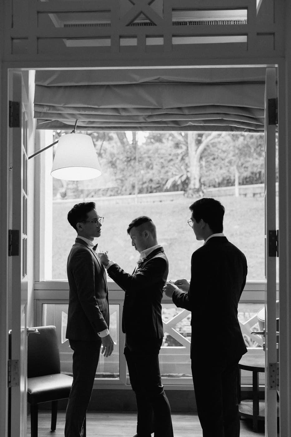 Black and white image of formal preparation scene with three figures by window.
