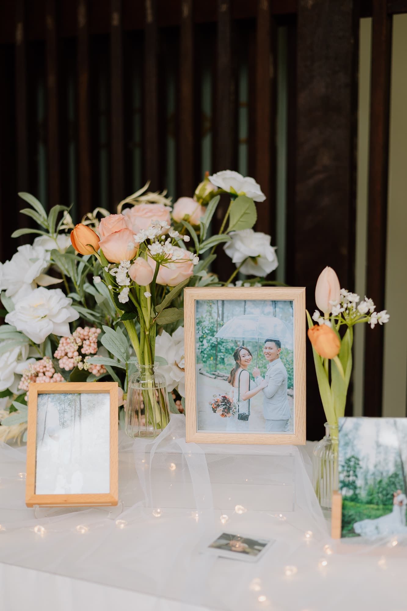 Wedding reception decor with framed photos and floral arrangements of roses and greenery on white table.
