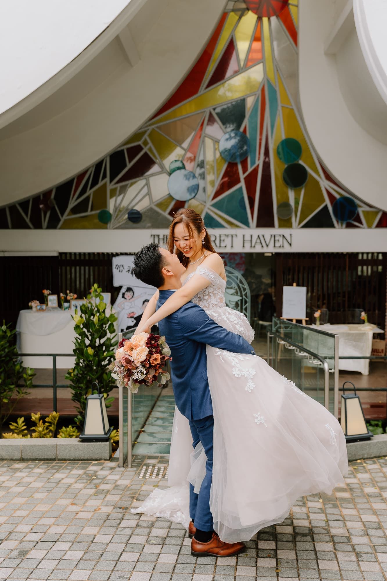 A couple shares a playful lift pose outside a venue decorated with colorful geometric art.