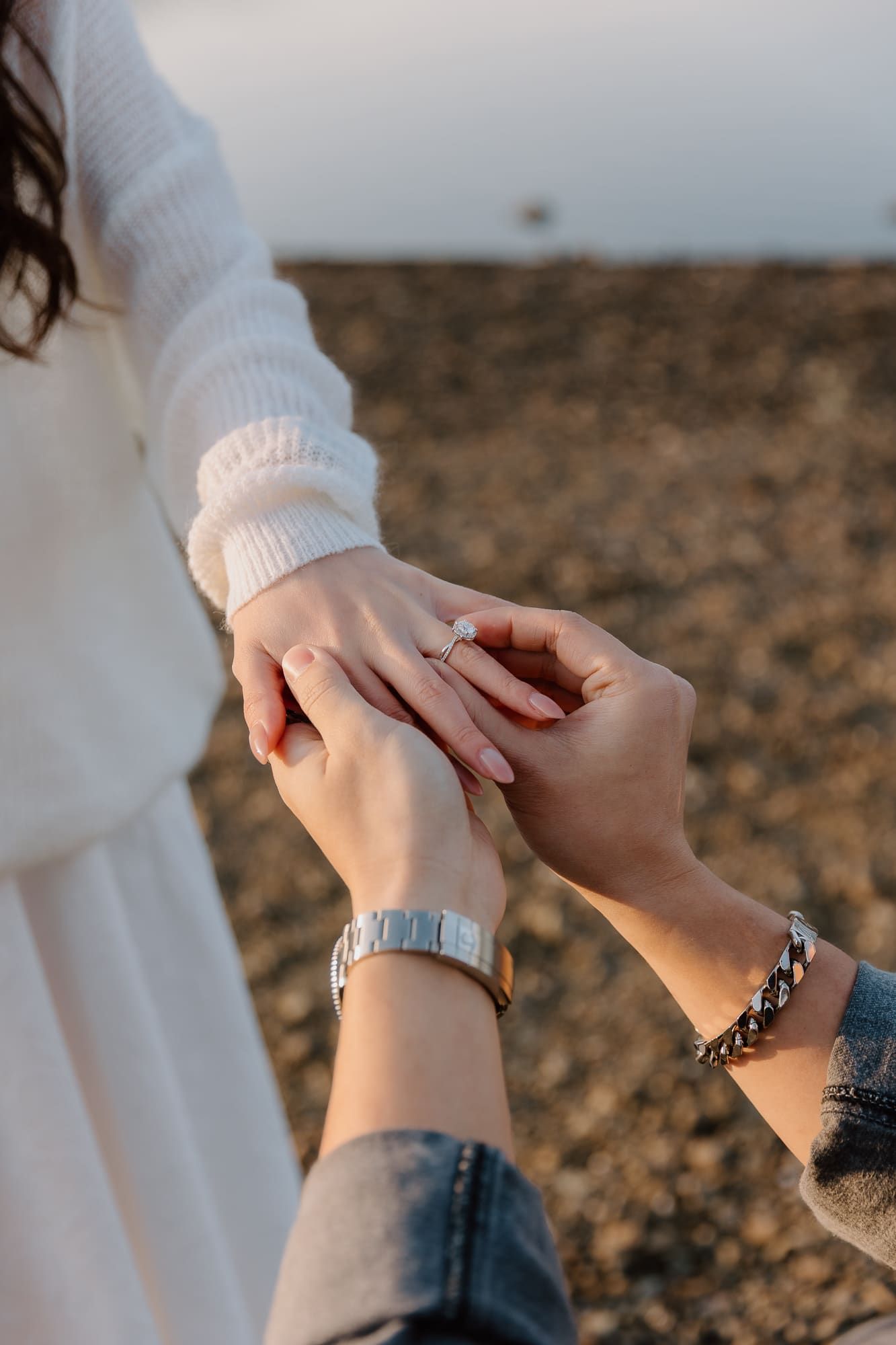 A person placing an engagement ring on their partner's finger outdoors near a rocky shore.