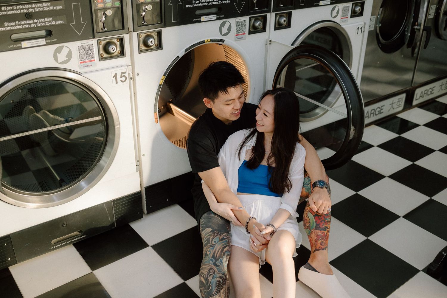 People loading clothes into washing machines at a laundromat with black and white checkered floor tiles.