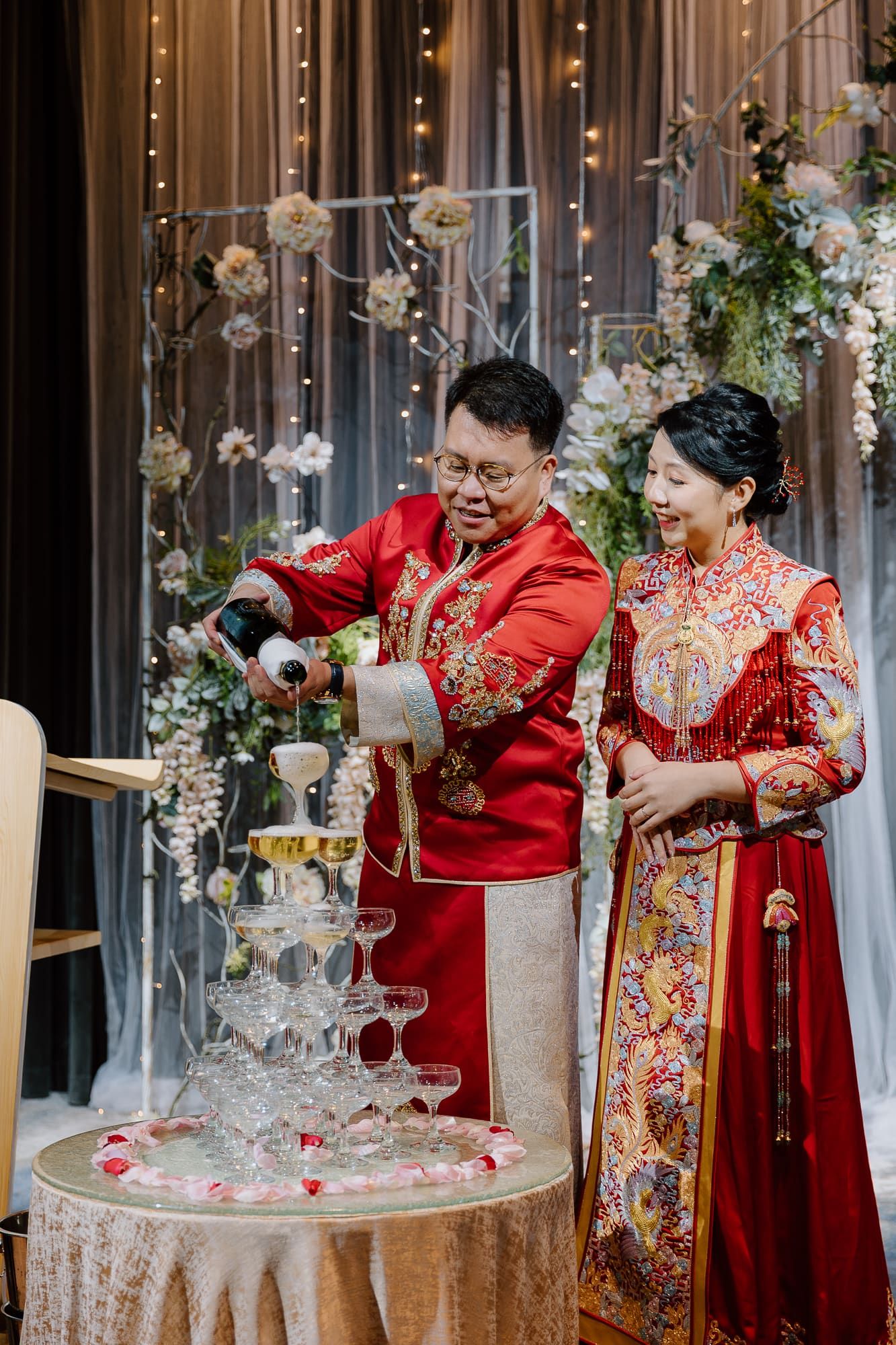A couple in traditional red Chinese wedding attire pour champagne into a tower of glasses at an elegant reception.