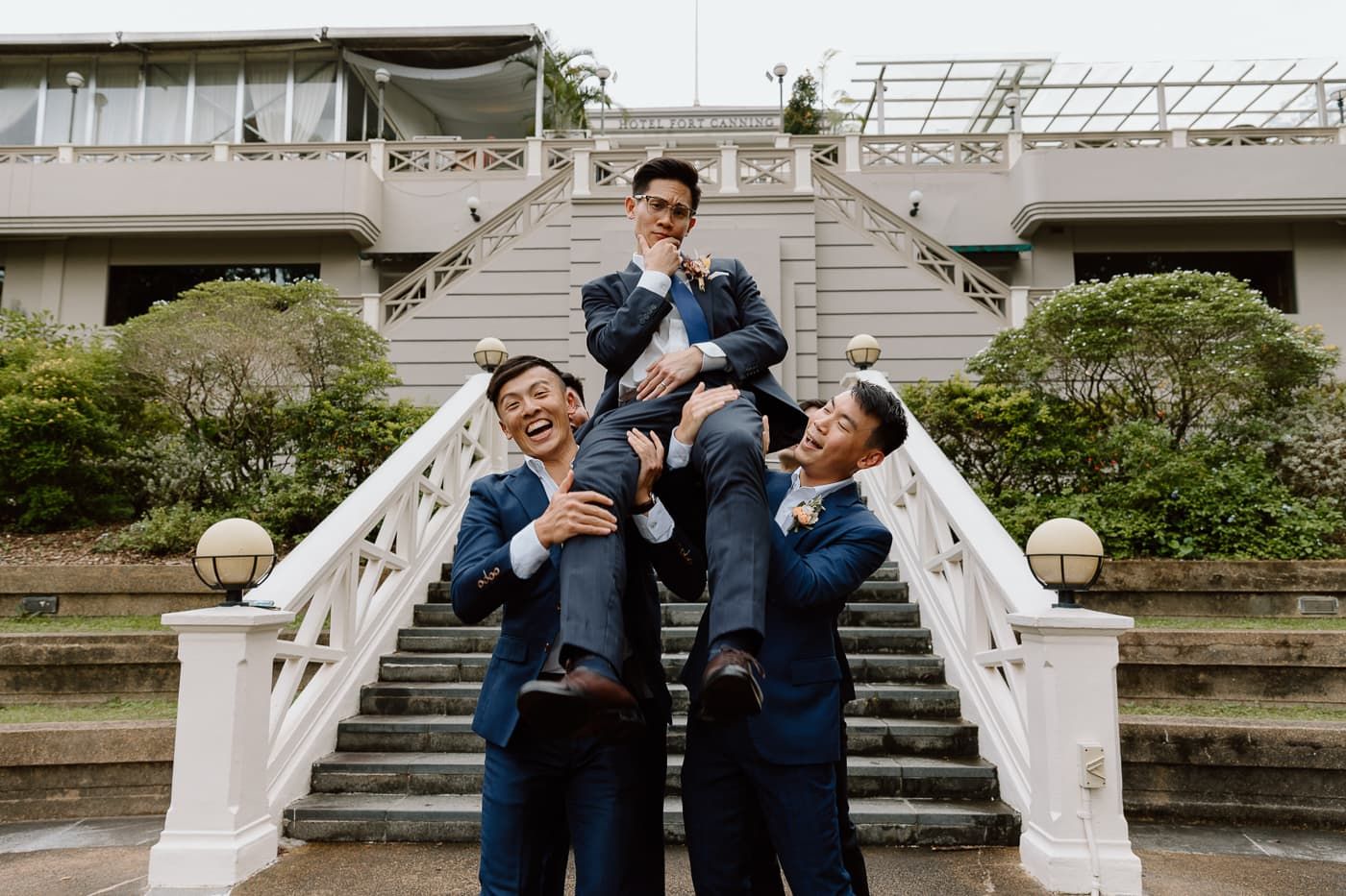 Playful groomsmen lift fellow member on grand white staircase during wedding photo session.