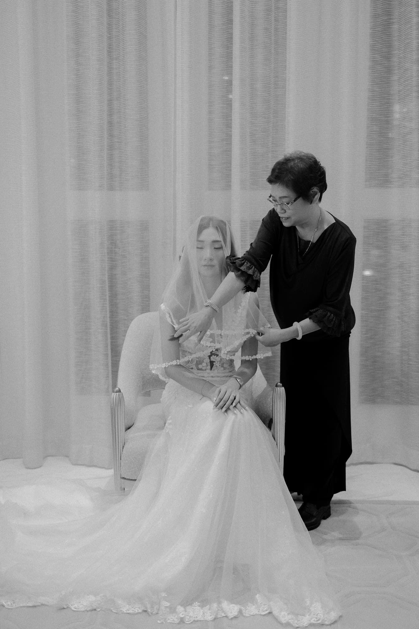 Black and white photo of bridal preparation scene in front of sheer curtains.