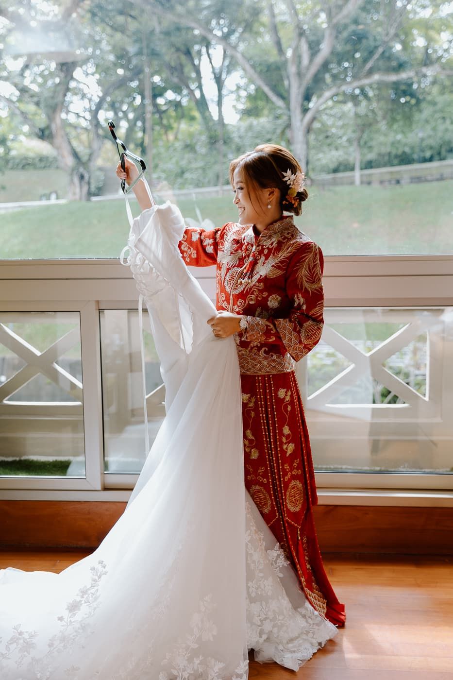 Person in red traditional dress holding white wedding gown near bright window.