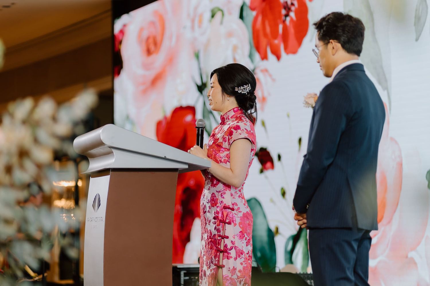 Wedding speakers at podium with red floral backdrop during reception celebration.