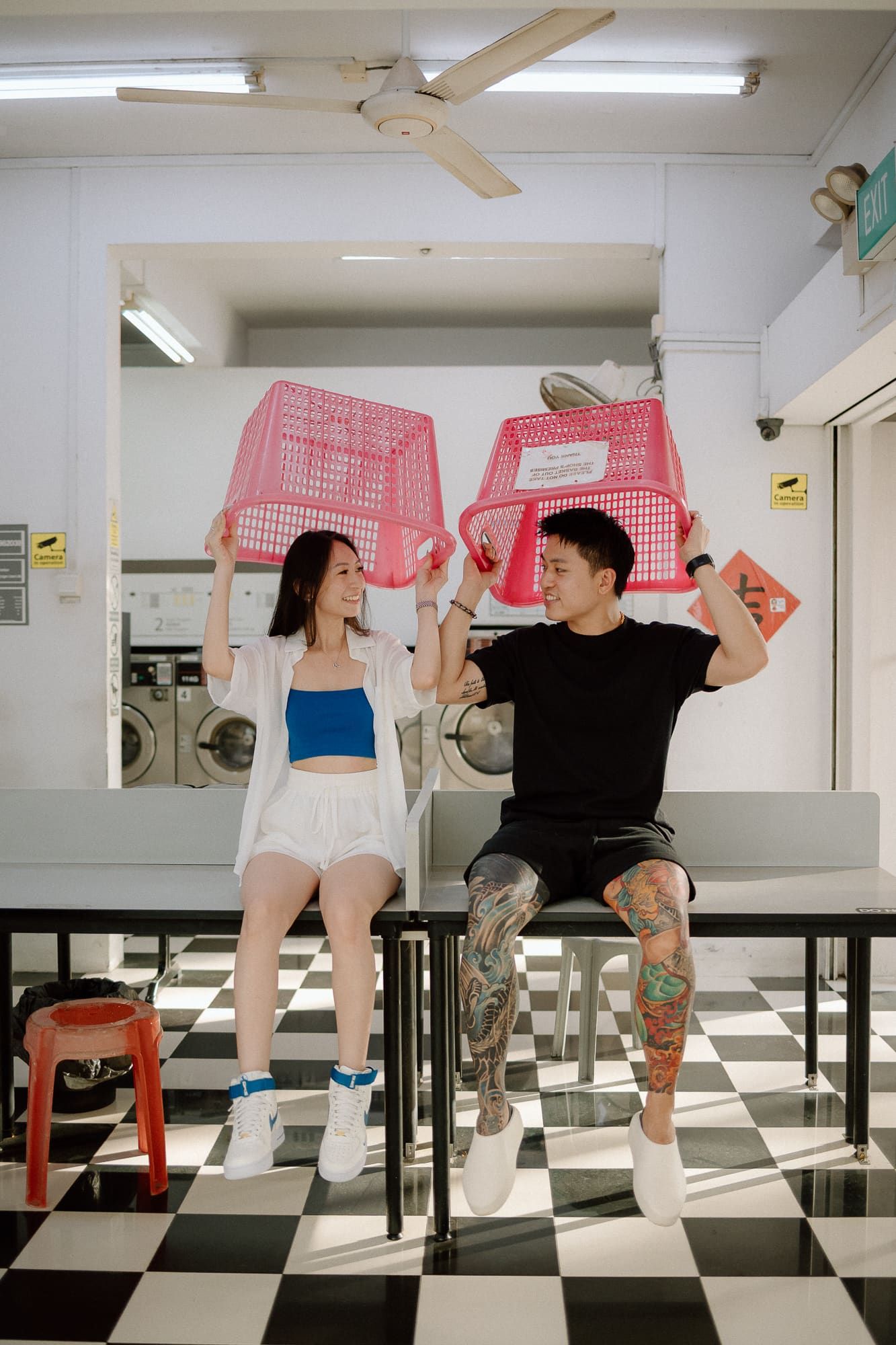 Two dancers perform a synchronized choreography on chairs in a black and white checkered room with pink props.
