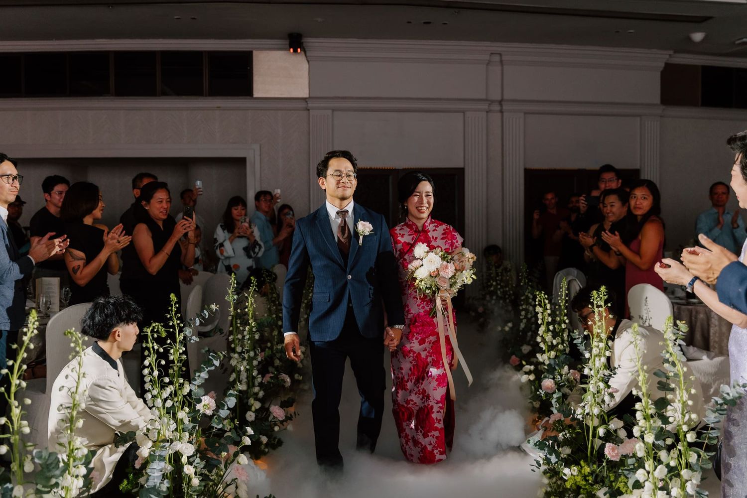 Wedding guests dance and celebrate during a reception with white floral arrangements in the background.