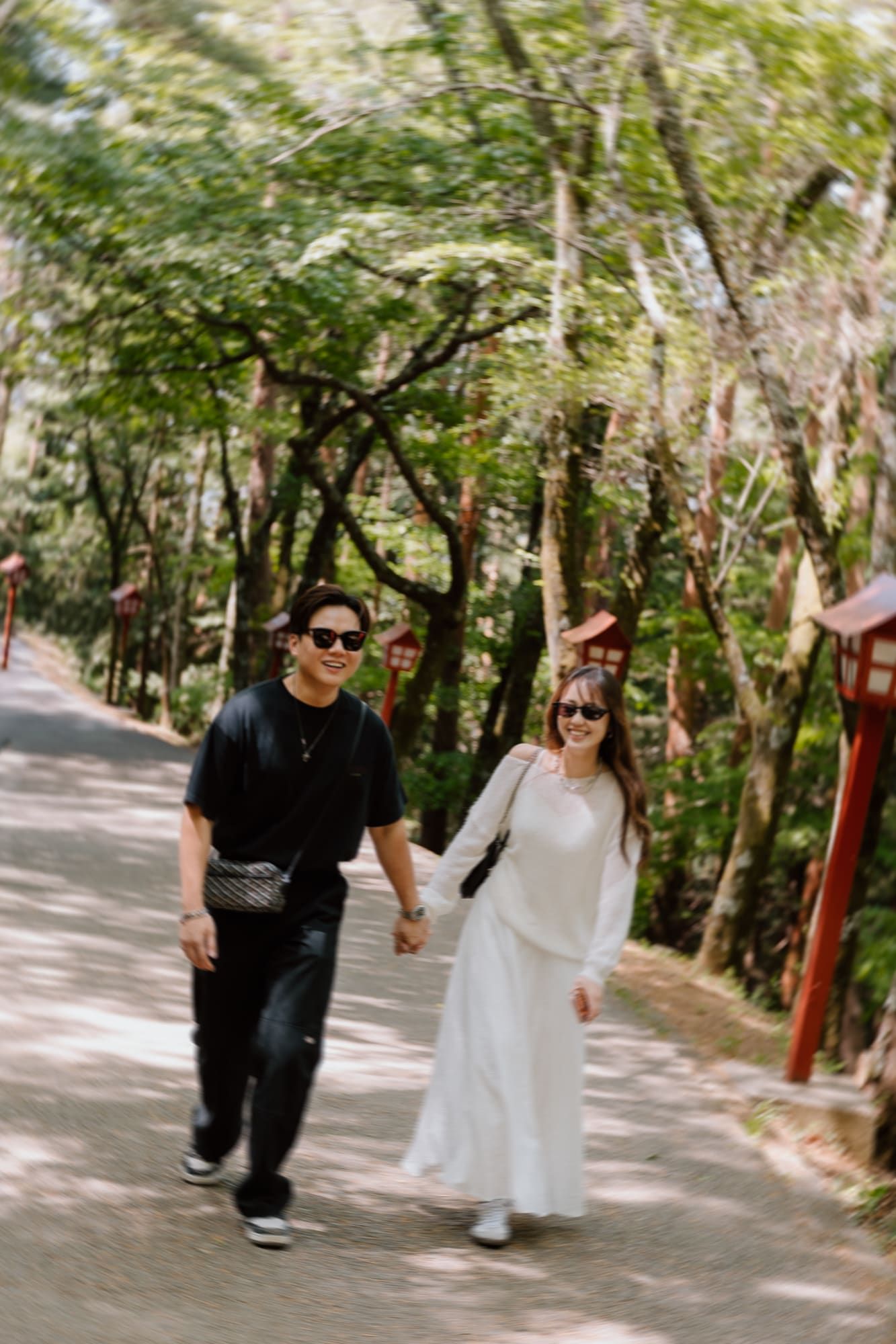 Couple holding hands walking on a tree-lined path, man in black outfit and woman in white dress wearing sunglasses.