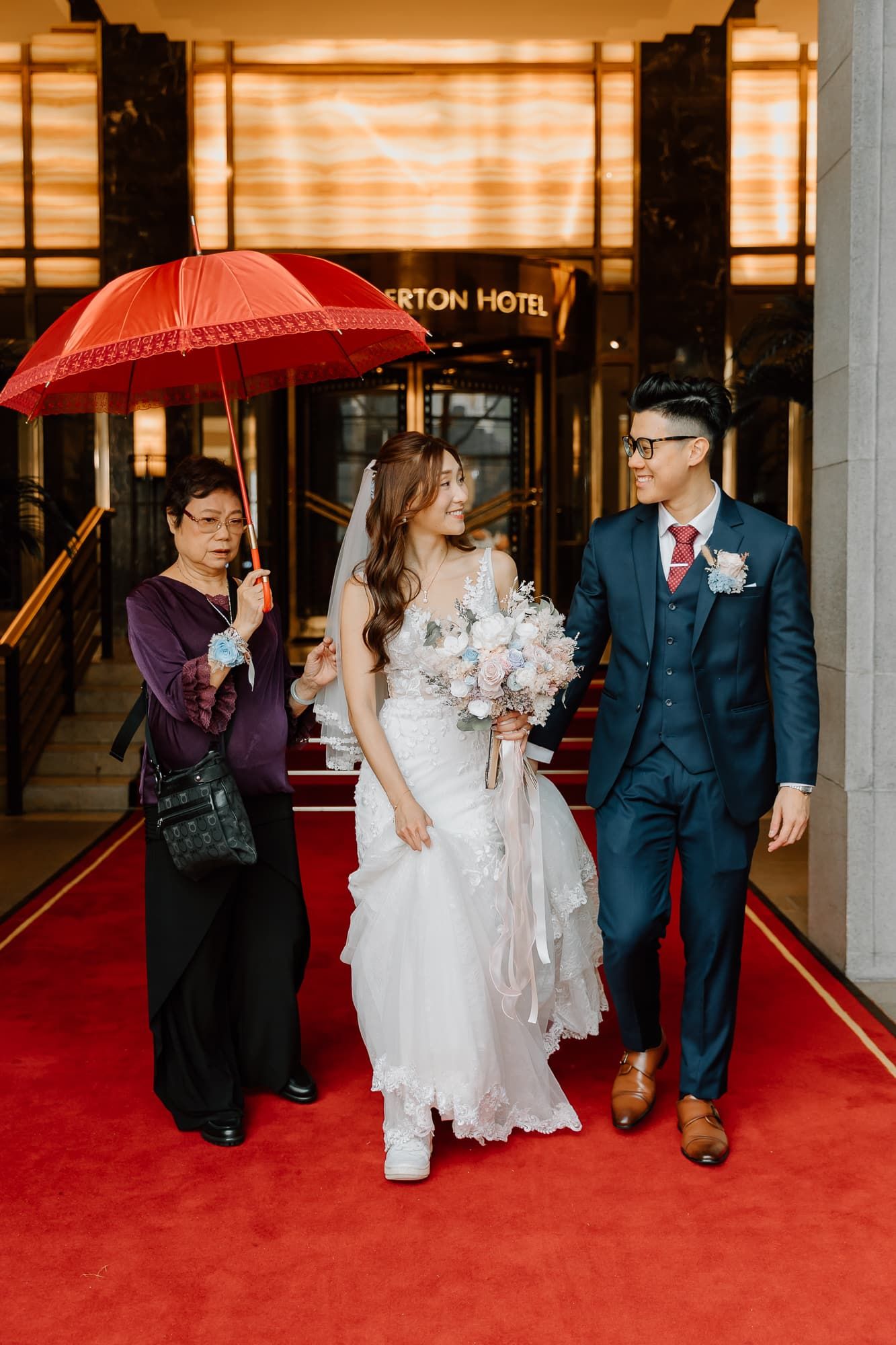 Wedding party walking on a red carpet under a red umbrella outside a hotel entrance.
