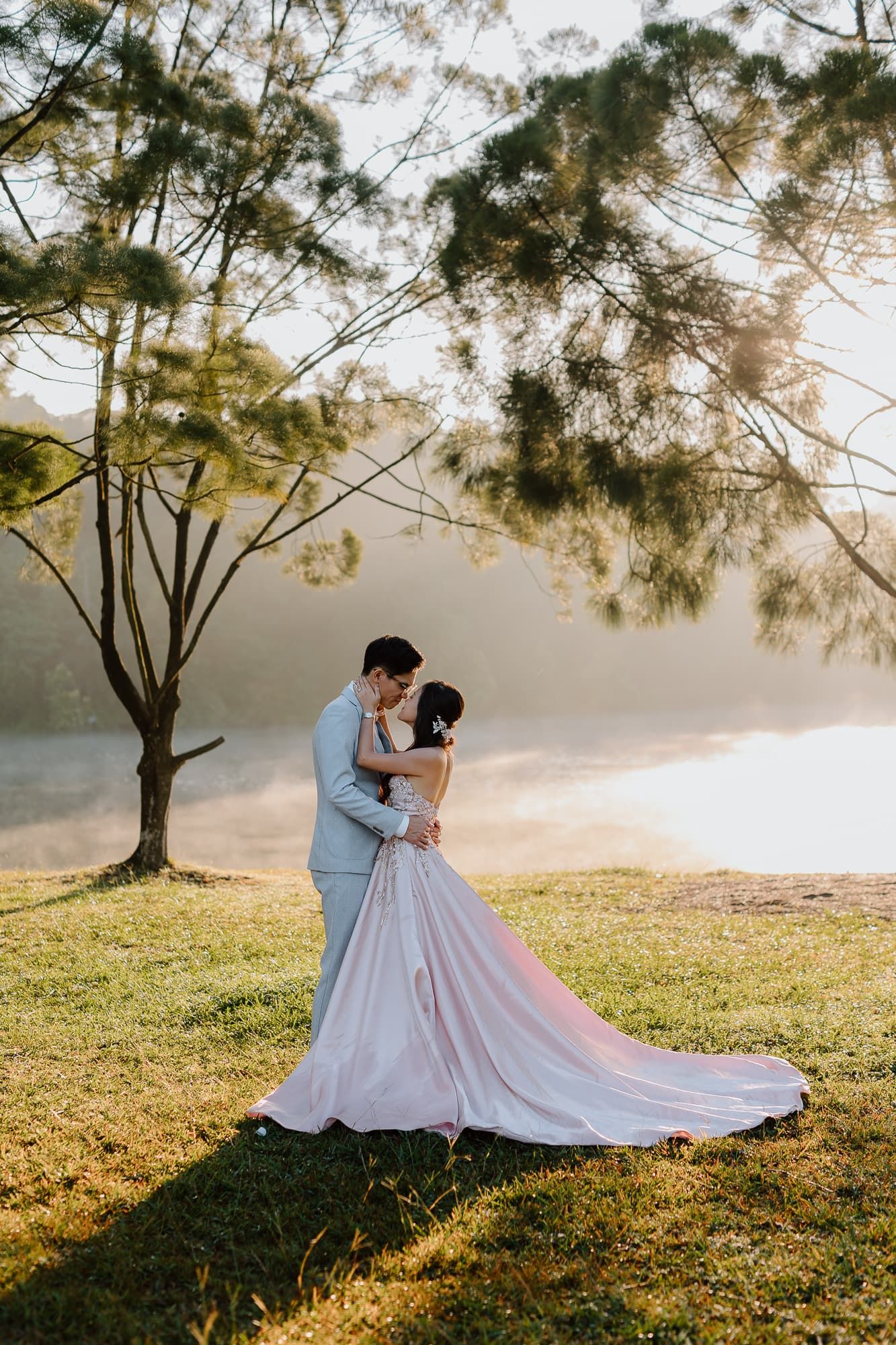 Couple embraces on lakeside hill during golden hour with dramatic lighting.