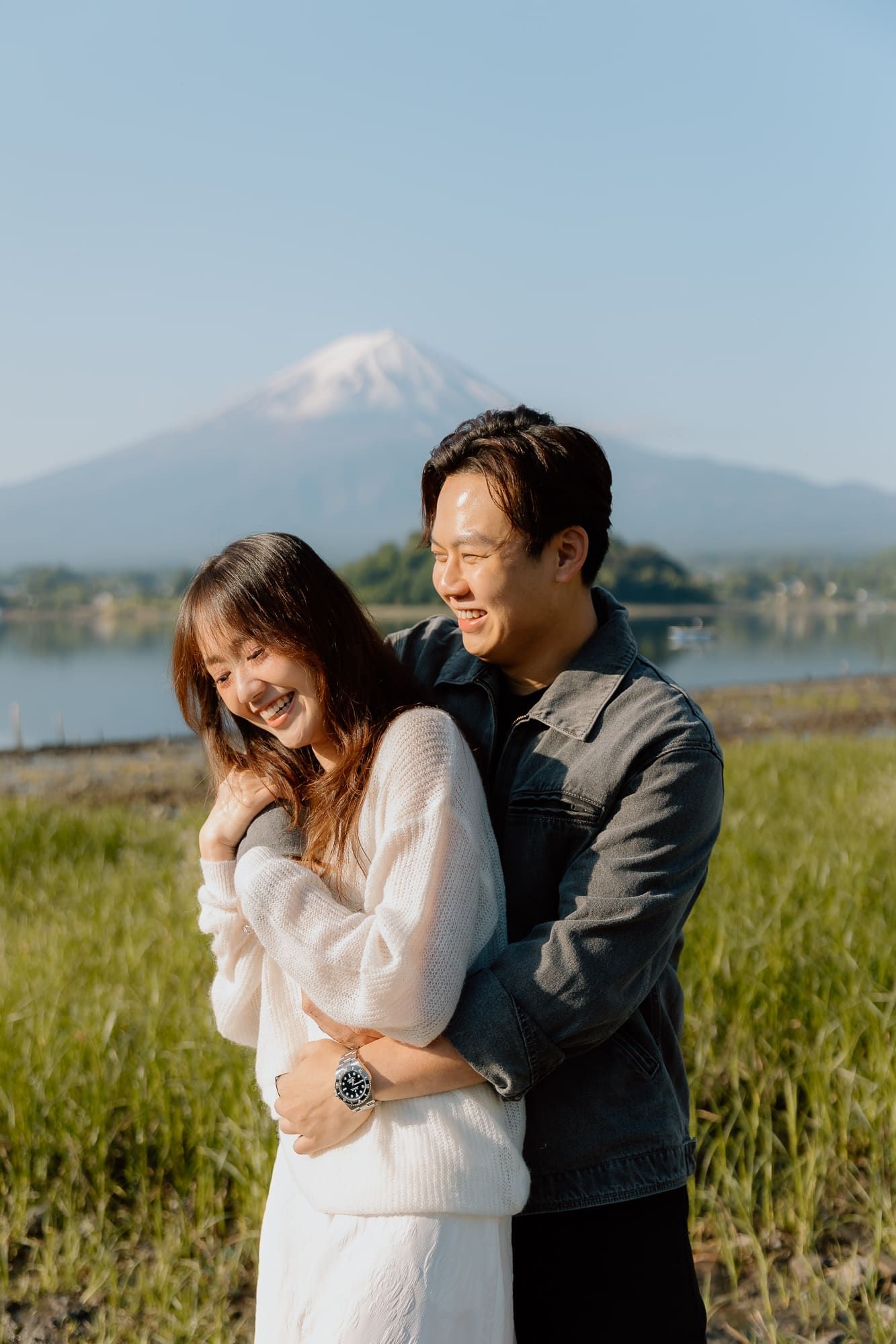 Happy couple embracing and laughing with Mount Fuji and a lake in the background on a clear day.