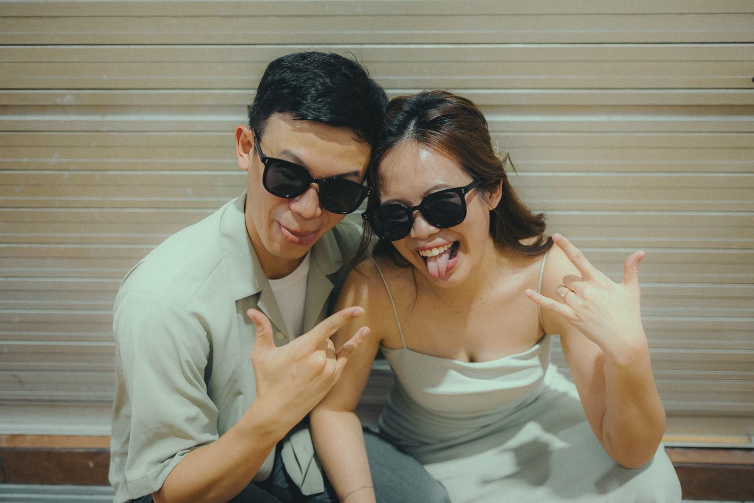 A couple wearing sunglasses making peace signs and smiling against a wooden background.