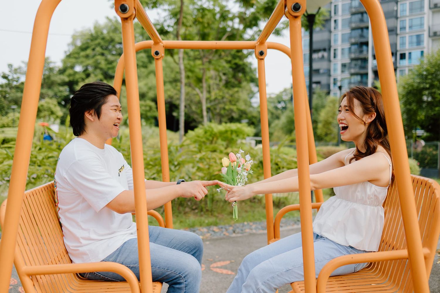 Couple sharing a playful moment while sitting on orange outdoor swings in a park setting.