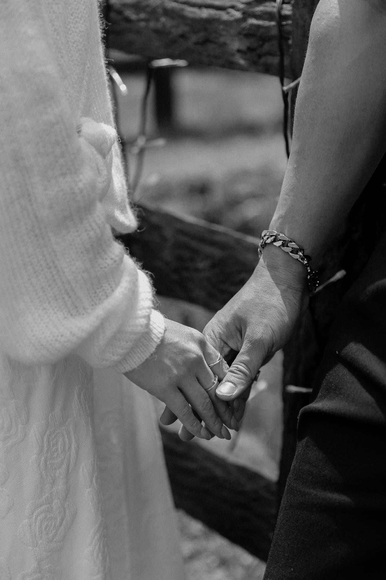 Black and white photo of a bride and groom holding hands, with the groom wearing a chain bracelet.