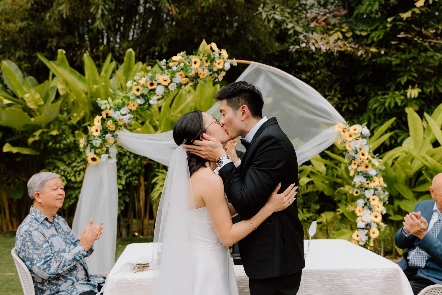 A couple shares their first kiss during an outdoor garden wedding ceremony under a floral arch.