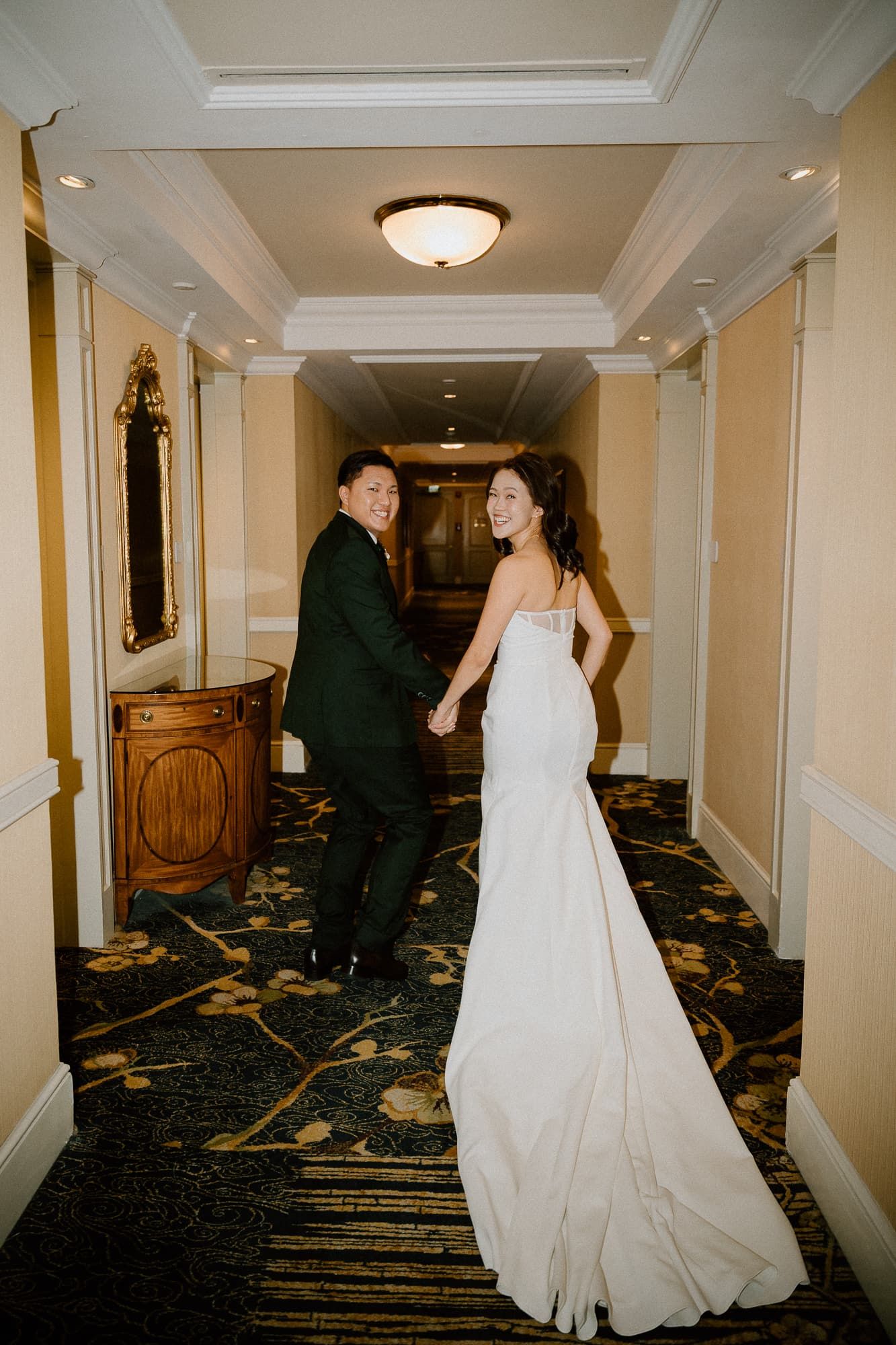 Wedding couple walking through elegant hotel hallway with classic crown molding and carpet.