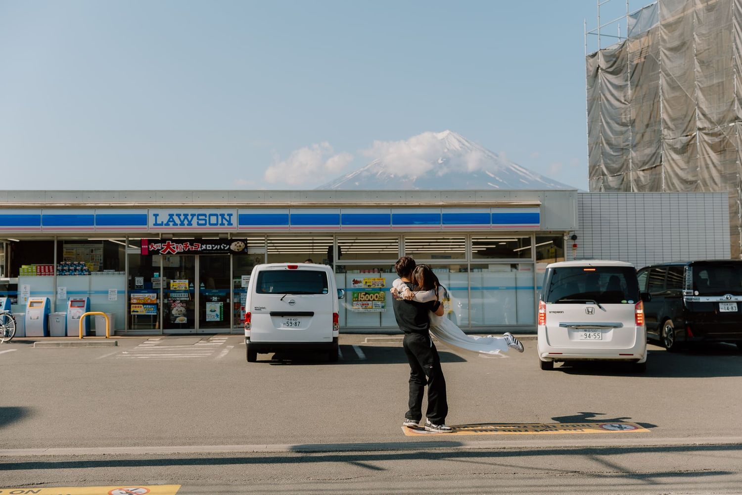Person photographing Mount Fuji behind a Lawson convenience store in Japan with vans parked in lot.