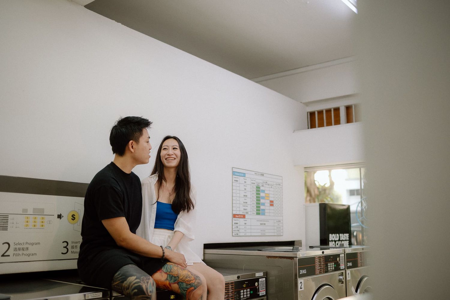 A couple sits together in a laundromat with industrial washing machines visible in the background.